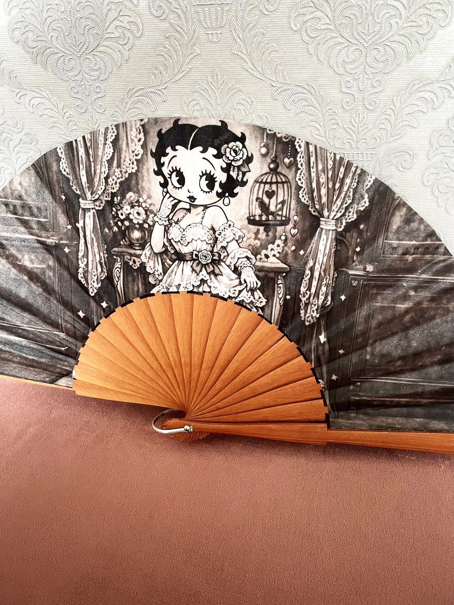 Détail du tissu exclusif d'un éventail en bois montrant l'illustration de Betty Boop pose rétro à côté d'une cage à oiseaux et de rideaux en dentelle.