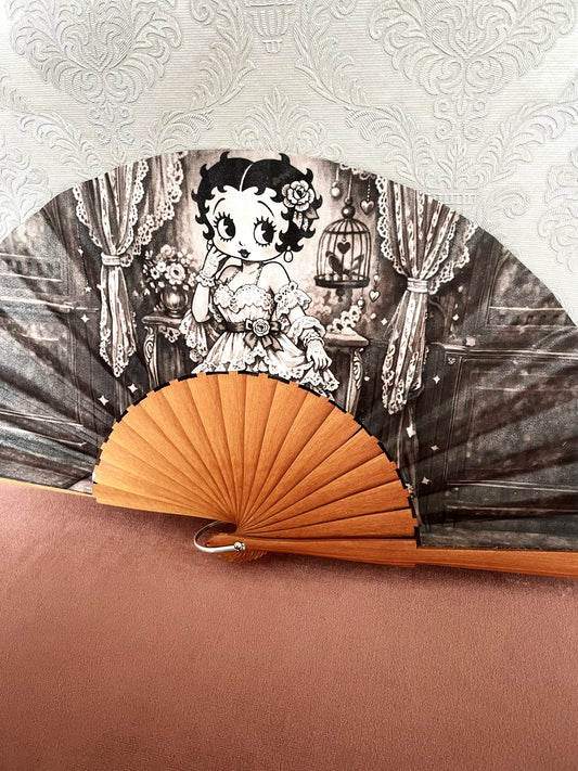 Détail du tissu exclusif d'un éventail en bois montrant l'illustration de Betty Boop pose rétro à côté d'une cage à oiseaux et de rideaux en dentelle.