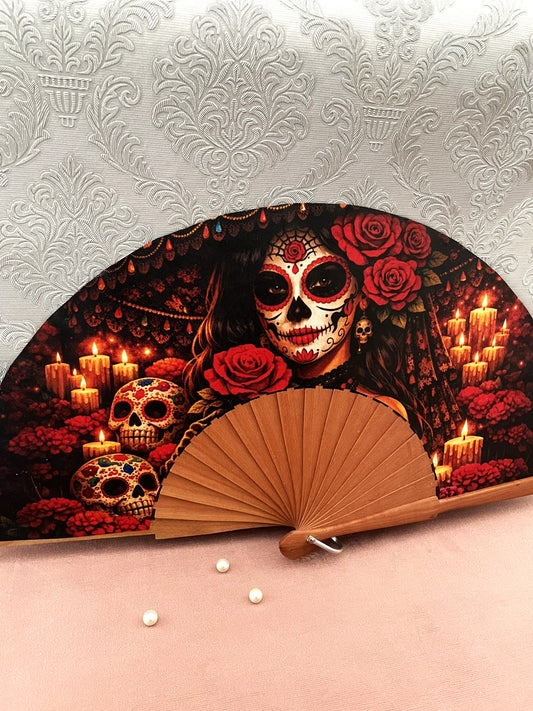Gros plan sur l'illustration du tissu exclusif d'un éventail fait main montrant le visage d'une Catrina maquillée, des roses rouges et des crânes décorés sous la lueur des bougies.