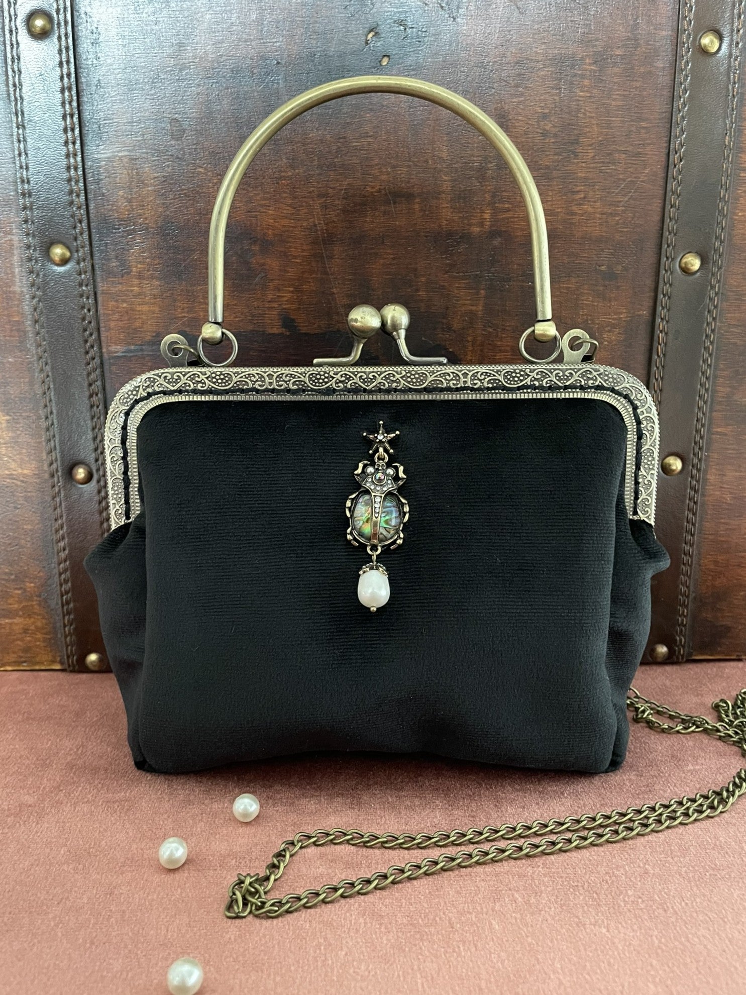 Gros plan sur le bijou scarabée serti et la perle suspendue sur le velours noir du sac.