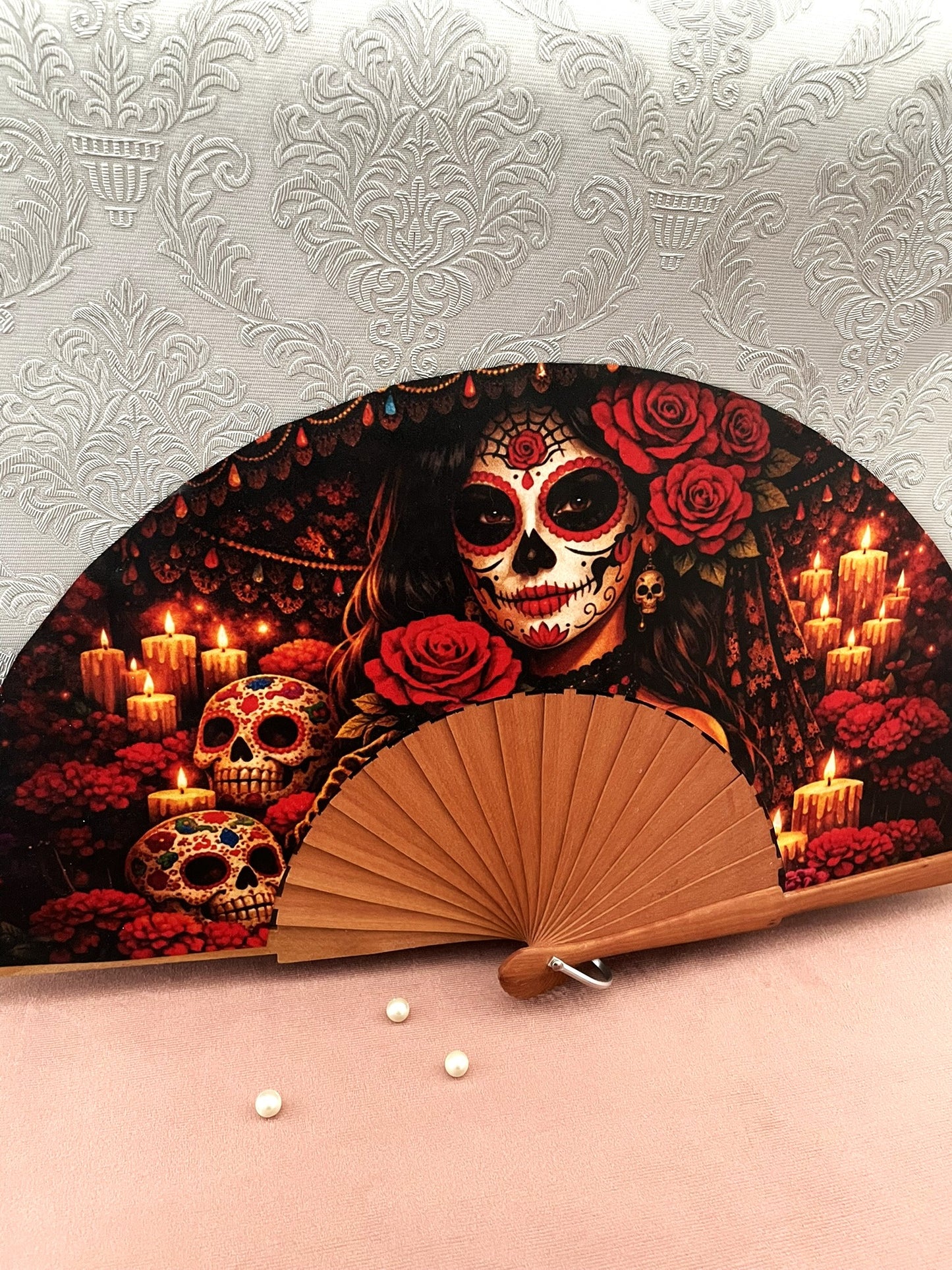Gros plan sur l'illustration du tissu exclusif d'un éventail fait main montrant le visage d'une Catrina maquillée, des roses rouges et des crânes décorés sous la lueur des bougies.