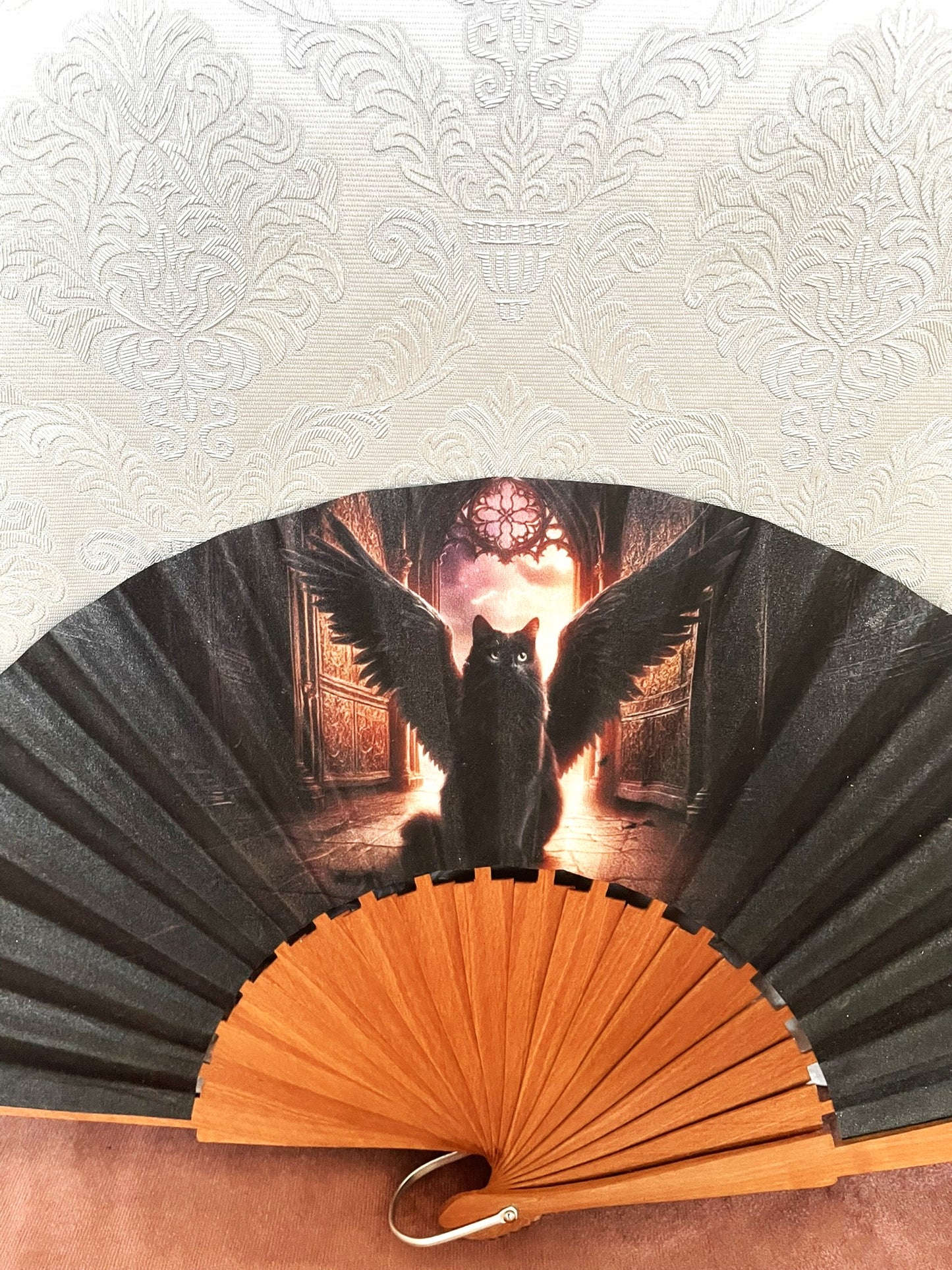 Gros plan sur le tissu exclusif d'un éventail fait main montrant l'illustration d'un chat noir avec de grandes ailes déployées devant une arche lumineuse.