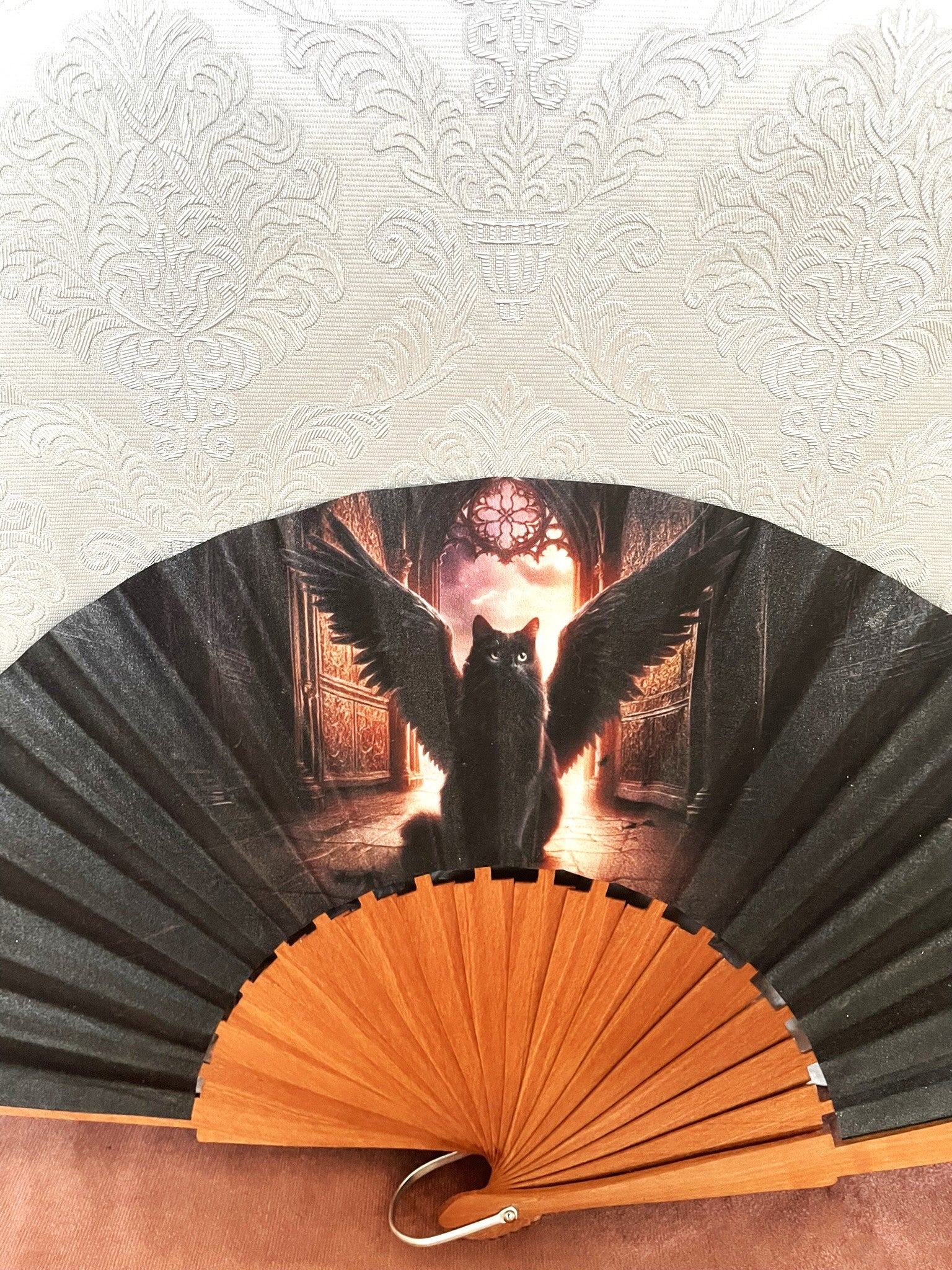Gros plan sur le tissu exclusif d'un éventail fait main montrant l'illustration d'un chat noir avec de grandes ailes déployées devant une arche lumineuse.