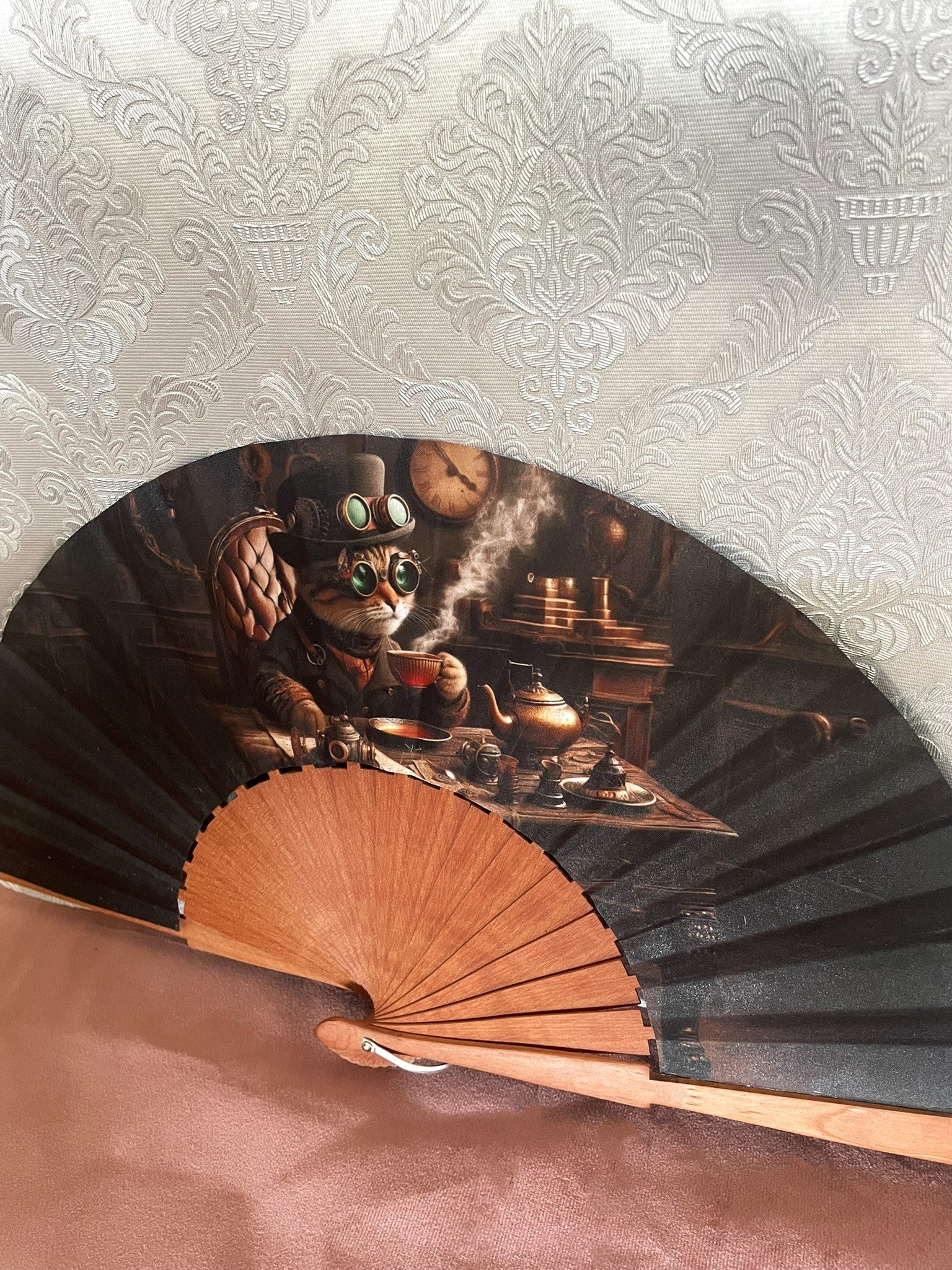 Gros plan sur le tissu exclusif d'un éventail fait main montrant un chat avec chapeau haut-de-forme, lunettes d'aviateur et tasse de thé dans un décor d'engrenages.