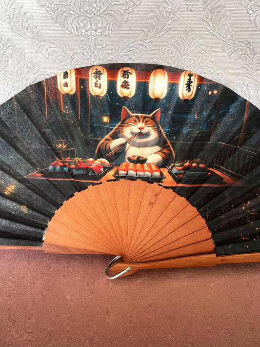 Détail du tissu exclusif d'un éventail en bois montrant l'illustration d'un chat souriant avec des baguettes devant un festin de sushis.