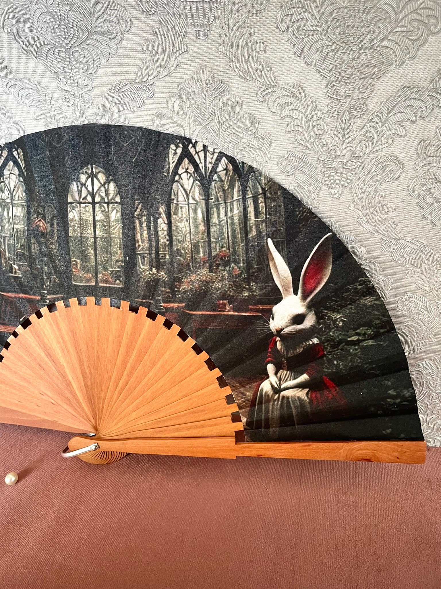 Détail du tissu exclusif d'un éventail en bois montrant un lapin blanc assis dans une robe bordeaux devant des vitraux et une verrerie gothique.