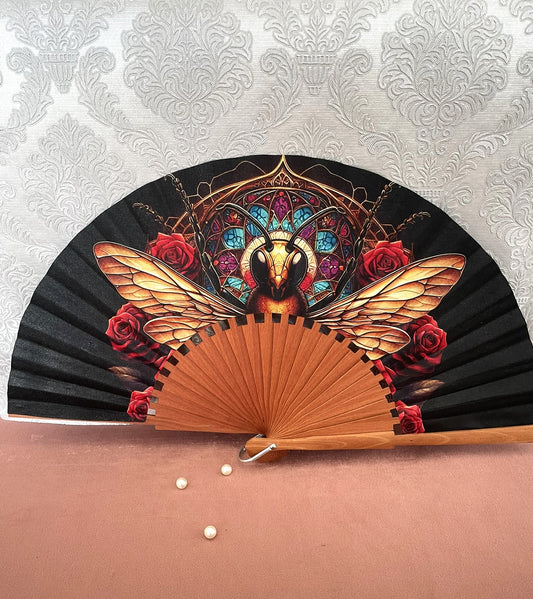 Éventail artisanal en bois fait main modèle abeille dorée devant un vitrail gothique et des roses rouges