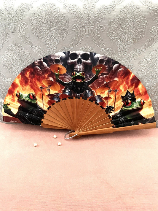 Éventail artisanal ouvert Le Comptoir de Suzanne en bois de fruitier avec tissu exclusif au motif humoristique de grenouilles rockeurs et batteur sur fond de flammes.