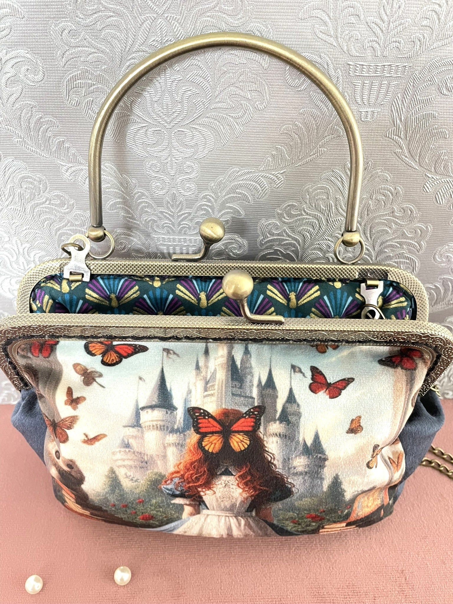 Pochette clutch féerique ouverte illustrant Alice de dos face à un château magique avec des papillons, révélant une doublure motif paon Art Déco.
