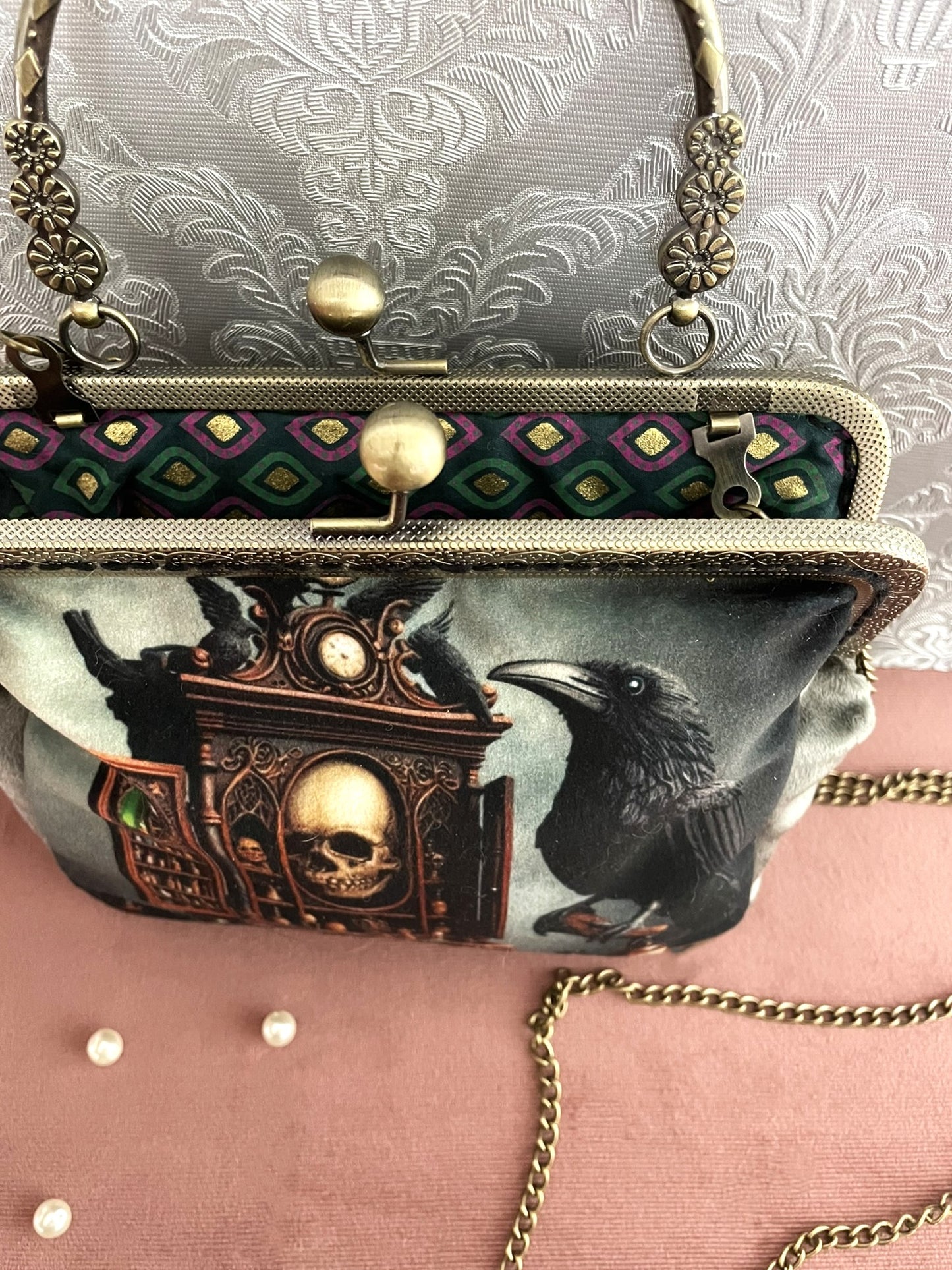 Vue du dessus de la pochette ouverte montrant sa doublure en tissu à motifs de losanges violets et dorés, son fermoir baiser et sa chaîne.