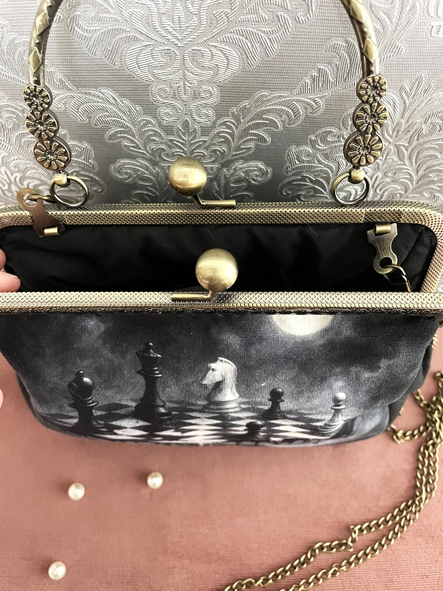 Vue du dessus de la pochette clutch échiquier ouverte montrant sa doublure en tissu noir uni, son fermoir clic-clac et l'attache de sa chaîne .