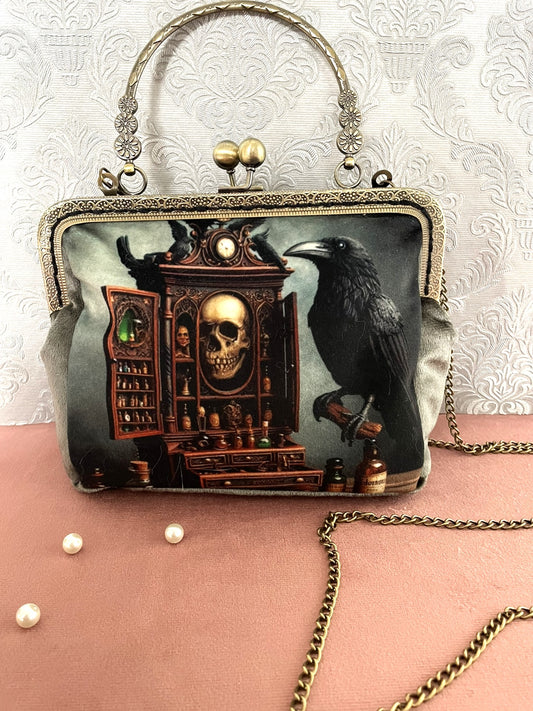 Pochette clutch gothique de face avec illustration de corbeau noir et meuble de sorcellerie avec crâne, poignée ciselée et chaîne.