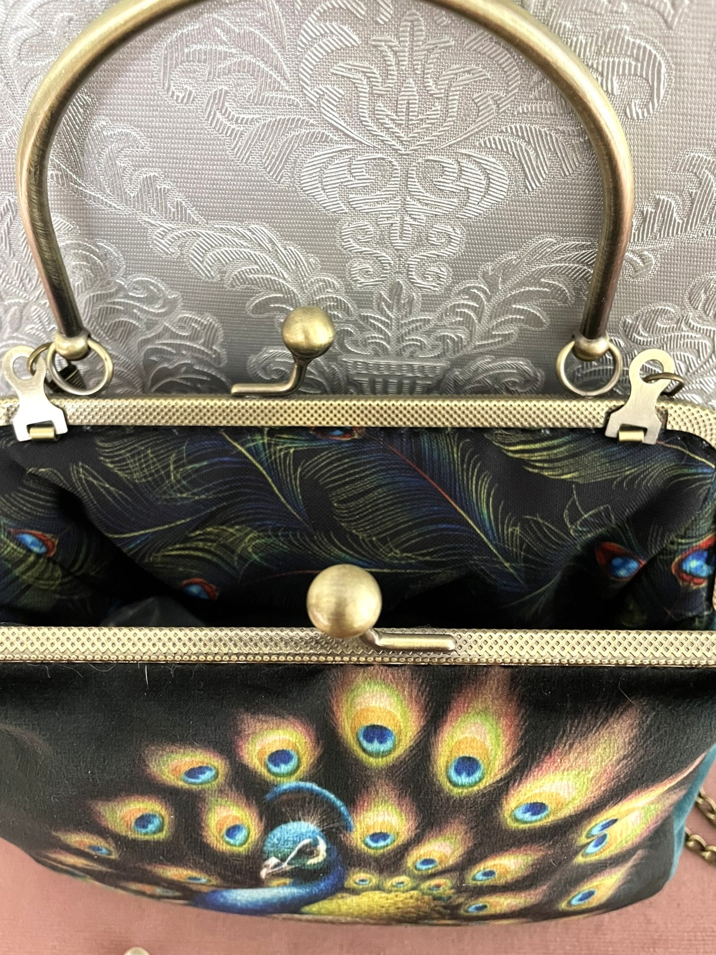 Vue du dessus de la pochette paon ouverte révélant le fermoir esprit vintage et sa doublure intérieure noire imprimée de plumes de paon bleues et vertes.