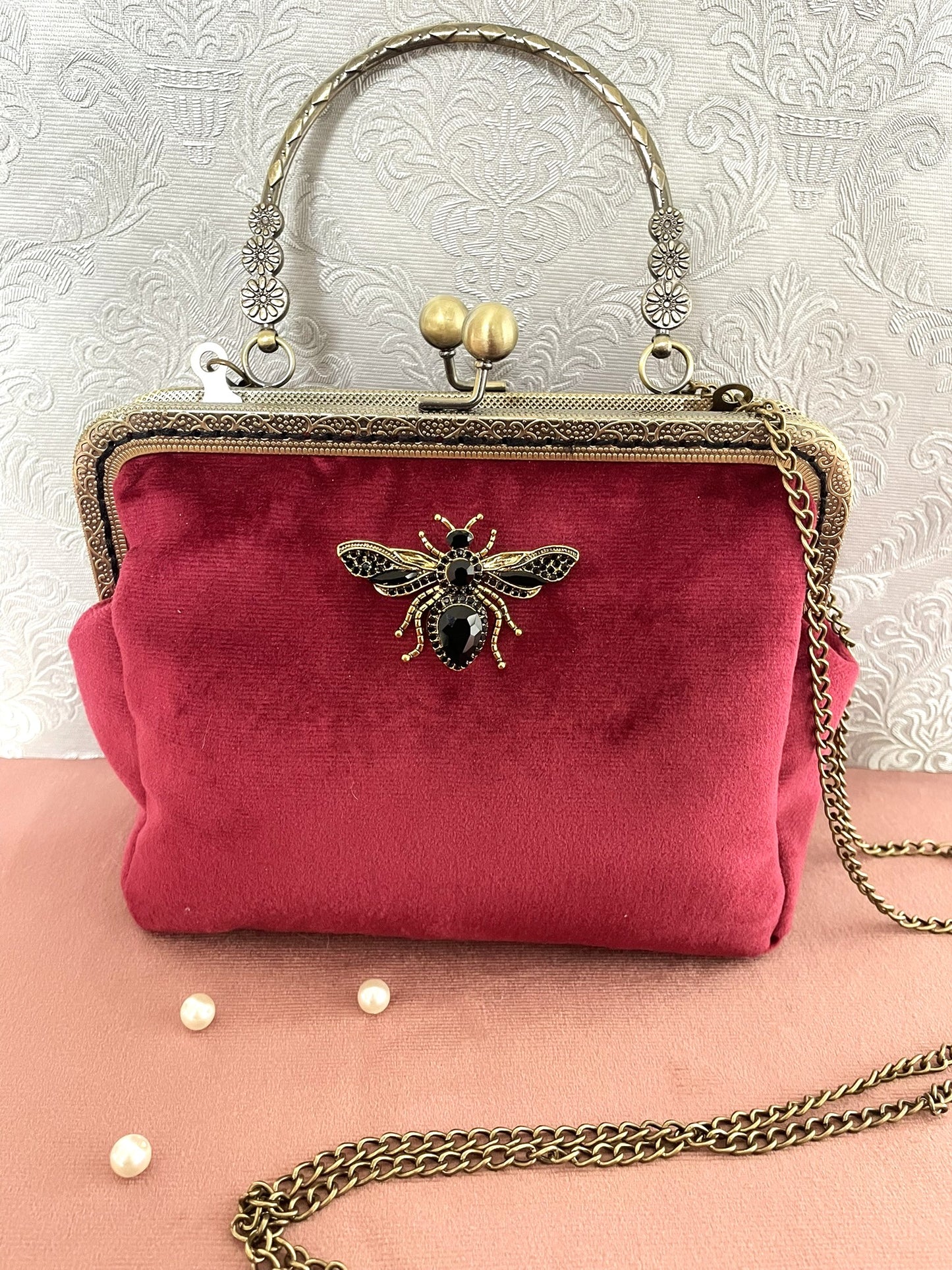 Pochette clutch de face en velours  bordeaux ornée d'une broche abeille en strass noirs et dorés, poignée ciselée et chaîne