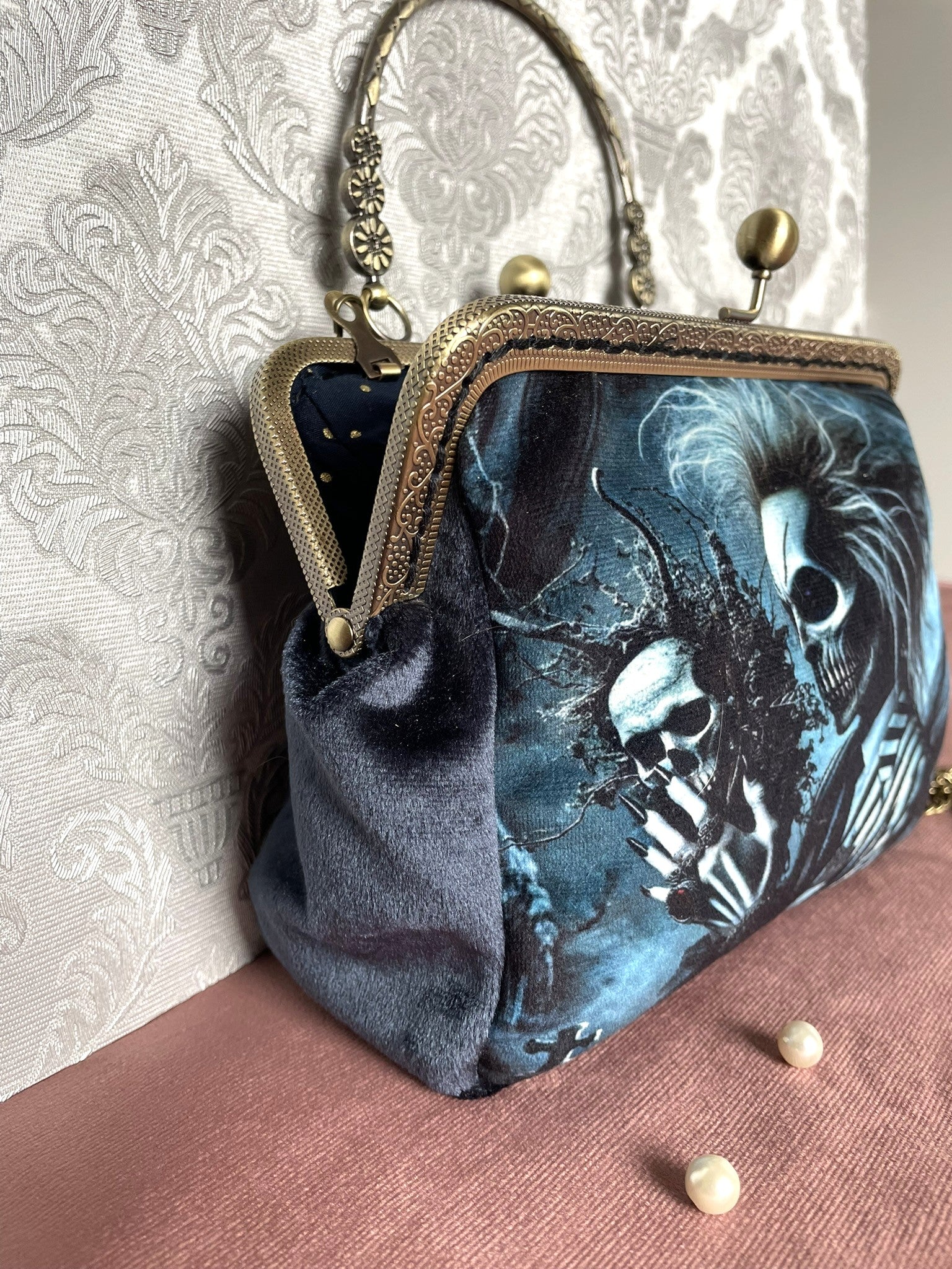 Gros plan latéral d'une pochette clutch gothique en velours bleu nuit avec illustration de crâne et fermoir clic-clac.