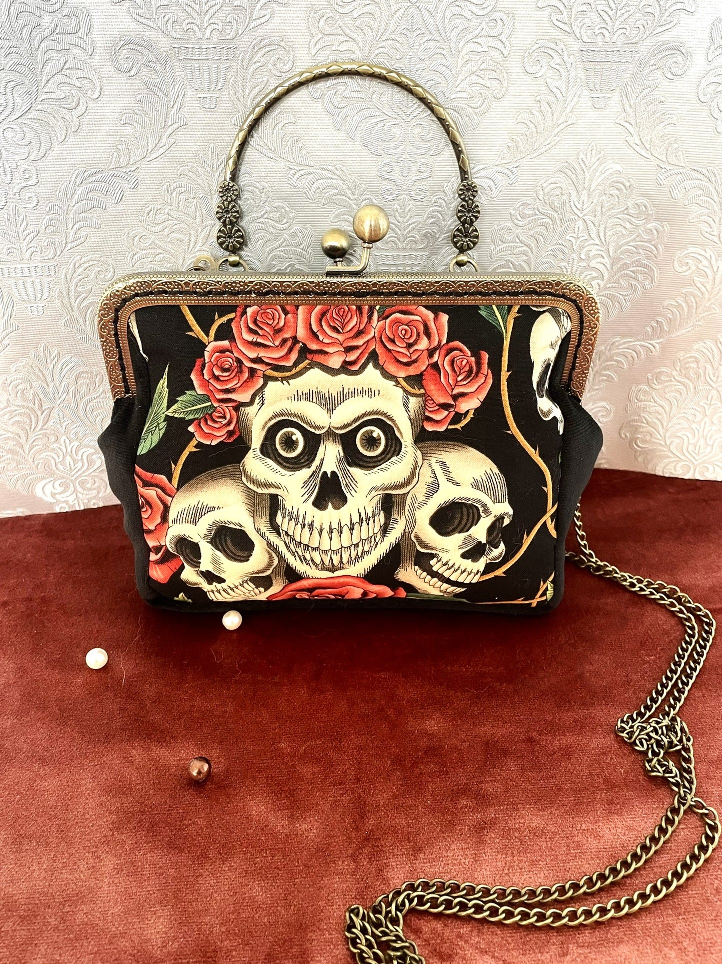 Sac à main rétro noir avec illustration de crânes (têtes de mort) et couronne de roses rouges, style gothique romantique.