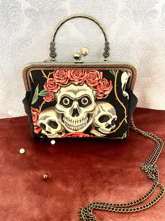 Sac à main rétro noir avec illustration de crânes (têtes de mort) et couronne de roses rouges, style gothique romantique.