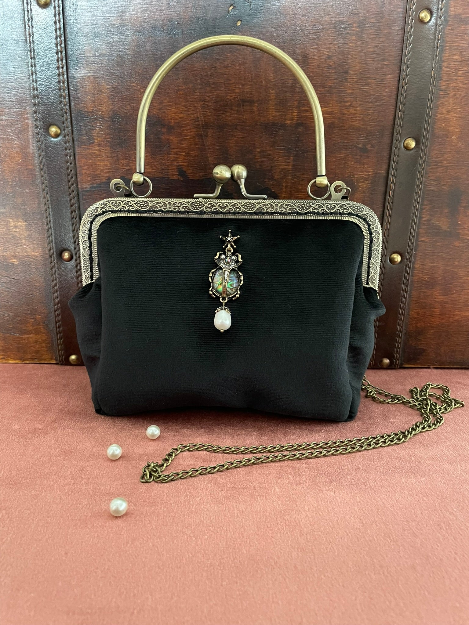 Sac à main vintage en velours noir avec bijou scarabée et anse bronze devant une malle ancienne.