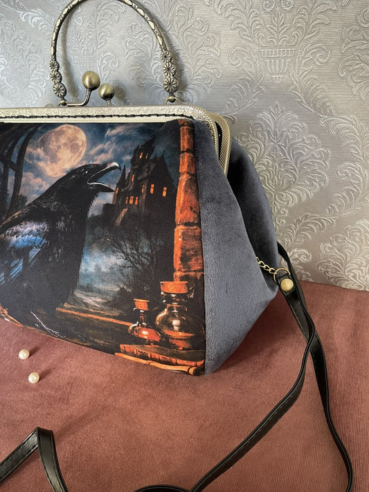 Détail de profil d'un sac à main rétro en velours gris bleu avec illustration d'un corbeau mystique et château gothique.